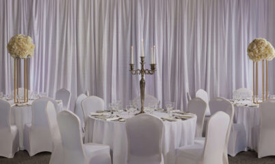 Elegant banquet hall set for guests with round tables, floral centrepieces, and candlelight.