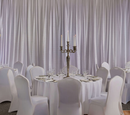 Elegant banquet hall set for guests with round tables, floral centrepieces, and candlelight.