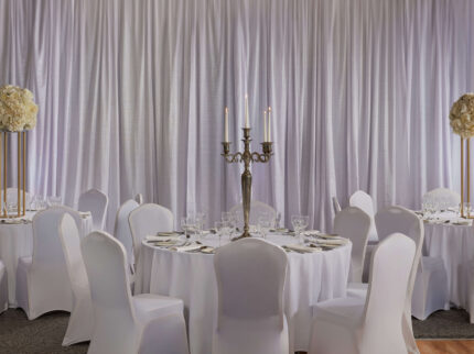 Elegant banquet hall set for guests with round tables, floral centrepieces, and candlelight.