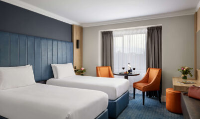 Cosy hotel room with twin beds, a table with wine, and welcoming decor for a relaxing stay.