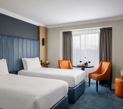 Cosy hotel room with twin beds, a table with wine, and welcoming decor for a relaxing stay.