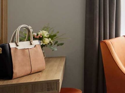 Elegant hotel room with warm lighting, a leather bag on the wooden desk, and an orange chair.