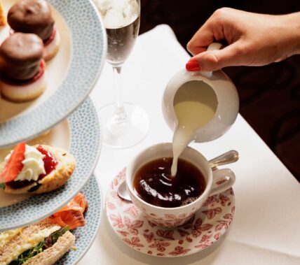 Hand pouring milk into tea with afternoon treats nearby.