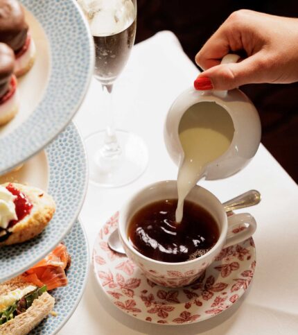 Hand pouring milk into tea with afternoon treats nearby.