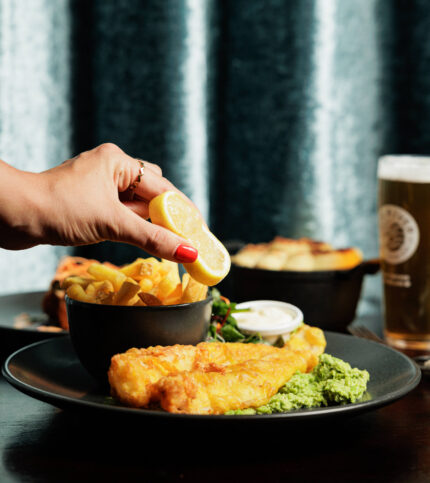 A hand squeezes lemon on fish and chips, with mushy peas and a pint in cosy setting.