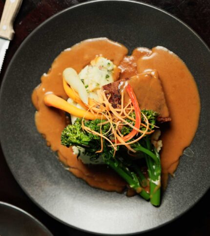 A gourmet steak dinner with vegetables and gravy, elegantly presented on a dark plate.