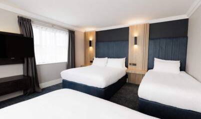Bright hotel room with three cosy beds, a large window, and warm lighting.