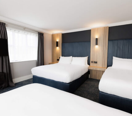 Bright hotel room with three cosy beds, a large window, and warm lighting.