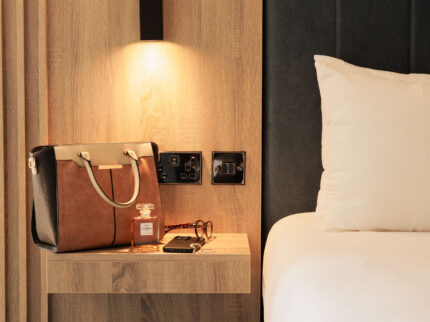 Cosy hotel room with modern decor and warm lighting; handbag and glasses on bedside table.