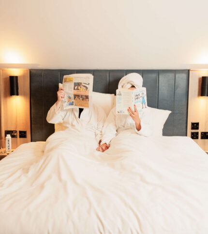 Couple in white bathrobes relaxing in a hotel bed, reading magazines.