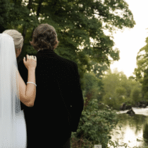 A couple in wedding attire admires a tranquil river setting surrounded by lush greenery.