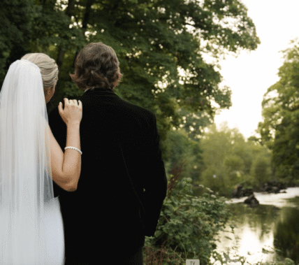 A couple in wedding attire admires a tranquil river setting surrounded by lush greenery.