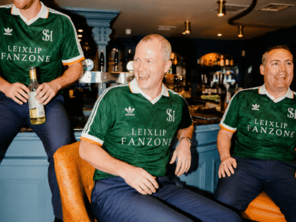 Three smiling men in matching green jerseys enjoy drinks at a cosy hotel bar.