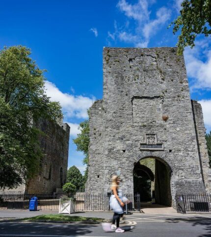 A couple strolls past a historic stone castle under a bright blue sky in Springfield, surrounded by lush trees.