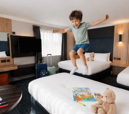 A joyful child jumps on a bed in a family-friendly hotel room with plush toys and activities.