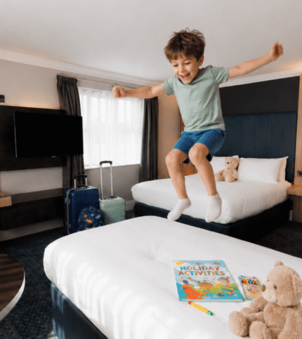 A joyful child jumps on a bed in a family-friendly hotel room with plush toys and activities.