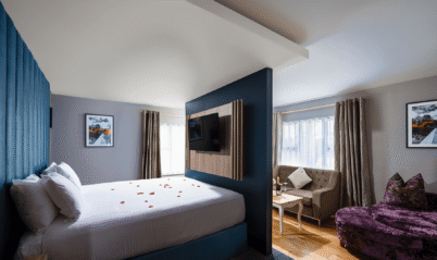 Modern hotel room with a cosy bed, rose petals, plush seating, and warm lighting.