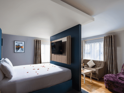 Modern hotel room with a cosy bed, rose petals, plush seating, and warm lighting.