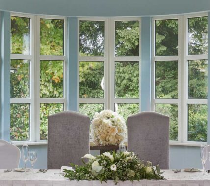 Elegant wedding reception table by large bay window with lush greenery outside.