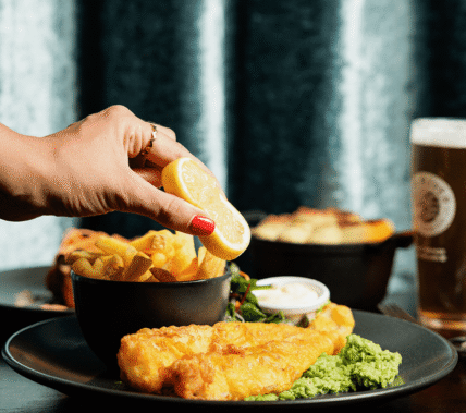 A hand squeezes lemon over fish and chips, with a pint of beer in a cosy setting.