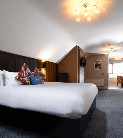 Spacious hotel room with double bed, desk, and suitcase, warmly lit for comfort.